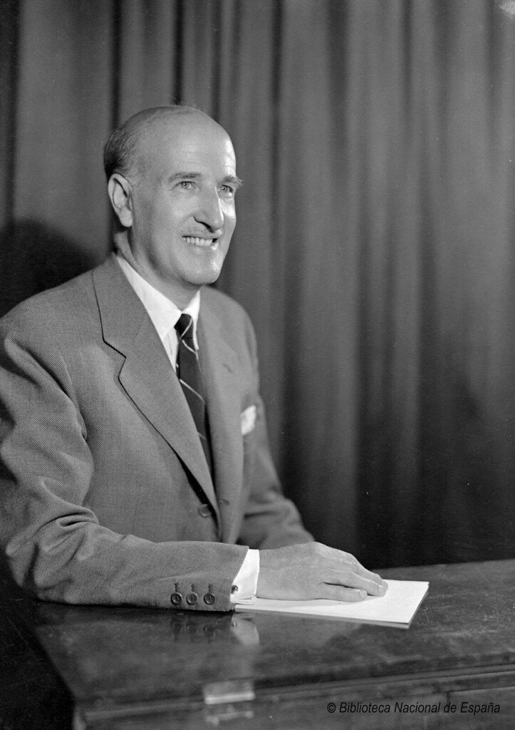 Imagen de Vicente Aleixandre en la que se le ve sonriente, sentado y sobre una mesa sostiene con su mano unos papeles