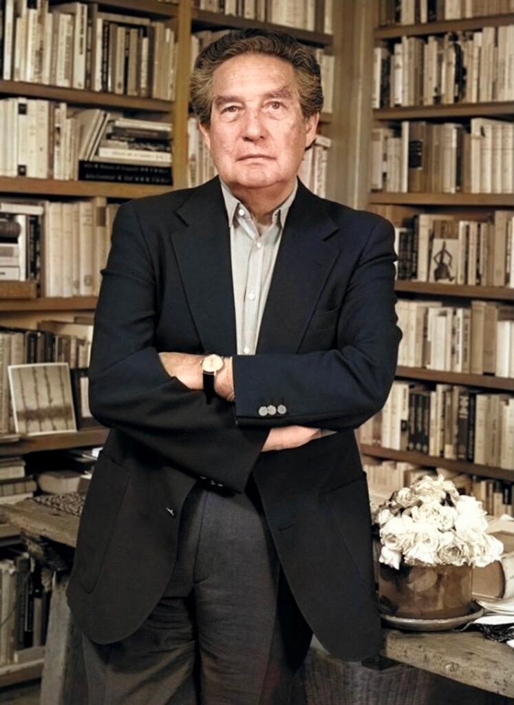 imagen de Octavio Paz, apoyado en una mesa con los brazos cruzados y detrás de él dos estanterías llenas de libros.