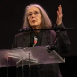 imagen de anne carson en la que parece de pie hablando tras un atril y un micrófono