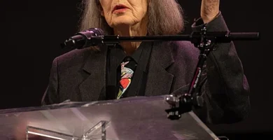 imagen de anne carson en la que parece de pie hablando tras un atril y un micrófono