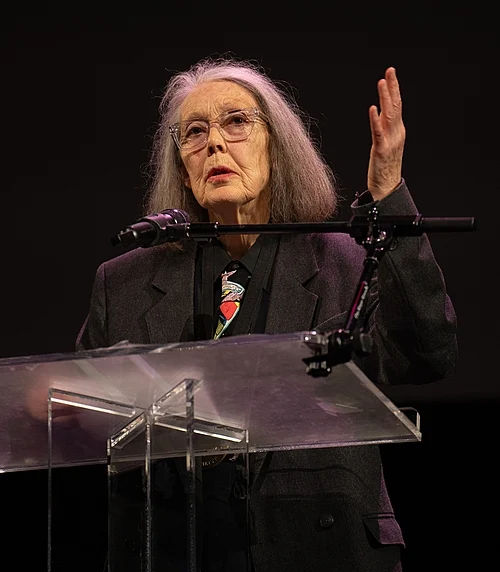 imagen de anne carson en la que parece de pie hablando tras un atril y un micrófono